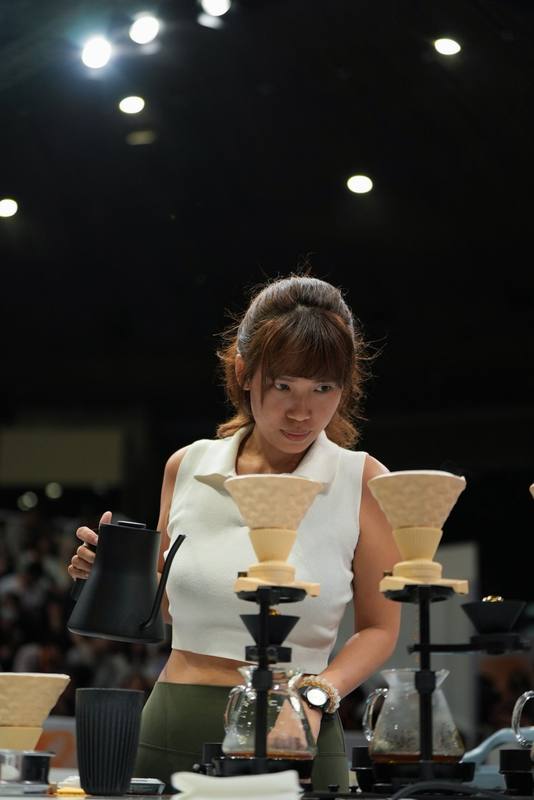 Photograph by Jeff Hann: A competitor pouring from a black kettle over two ceramic drippers, stage lights above