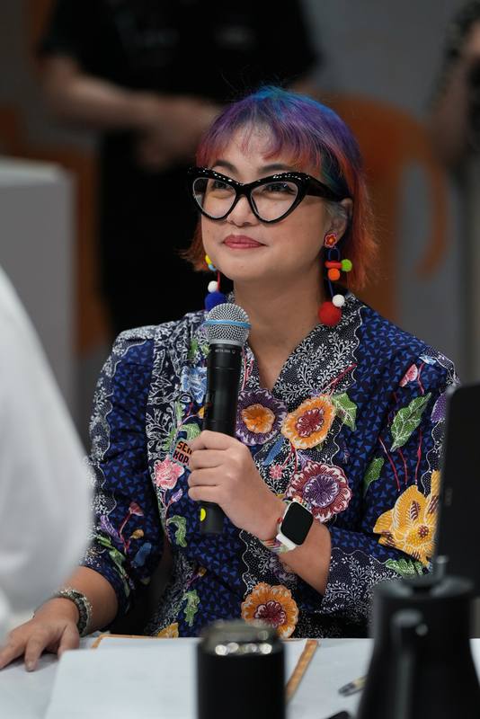 Photograph by Jeff Hann: A judge with rainbow hair, cat-eye glasses and a floral batik shirt, holding a microphone