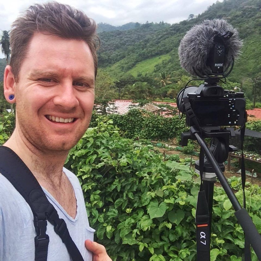 Jeff smiling beside his camera on a tripod, above a green hillside farm