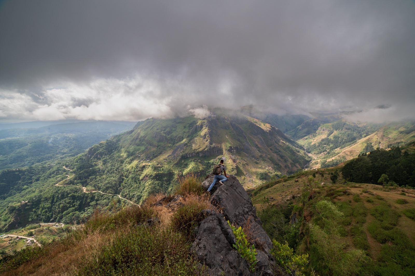 A man with a backpack climbing out along a rock ridge above a deep green valley, under low grey cloud