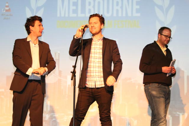 Three men on stage in front of a Melbourne Documentary Film Festival backdrop, one speaking into a microphone